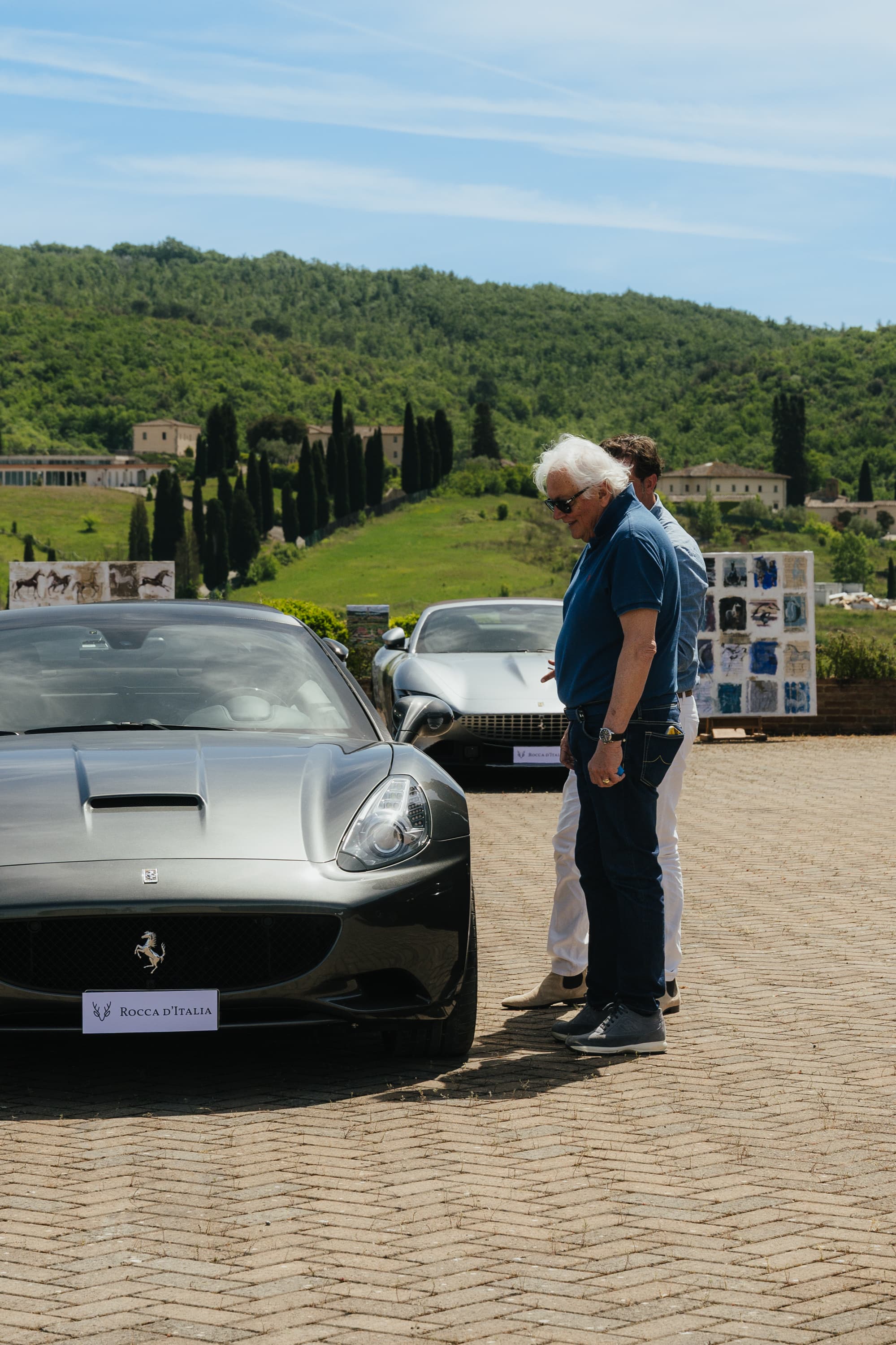 Ferrari grigia con targa Rocca d'Italia tra i cipressi toscani